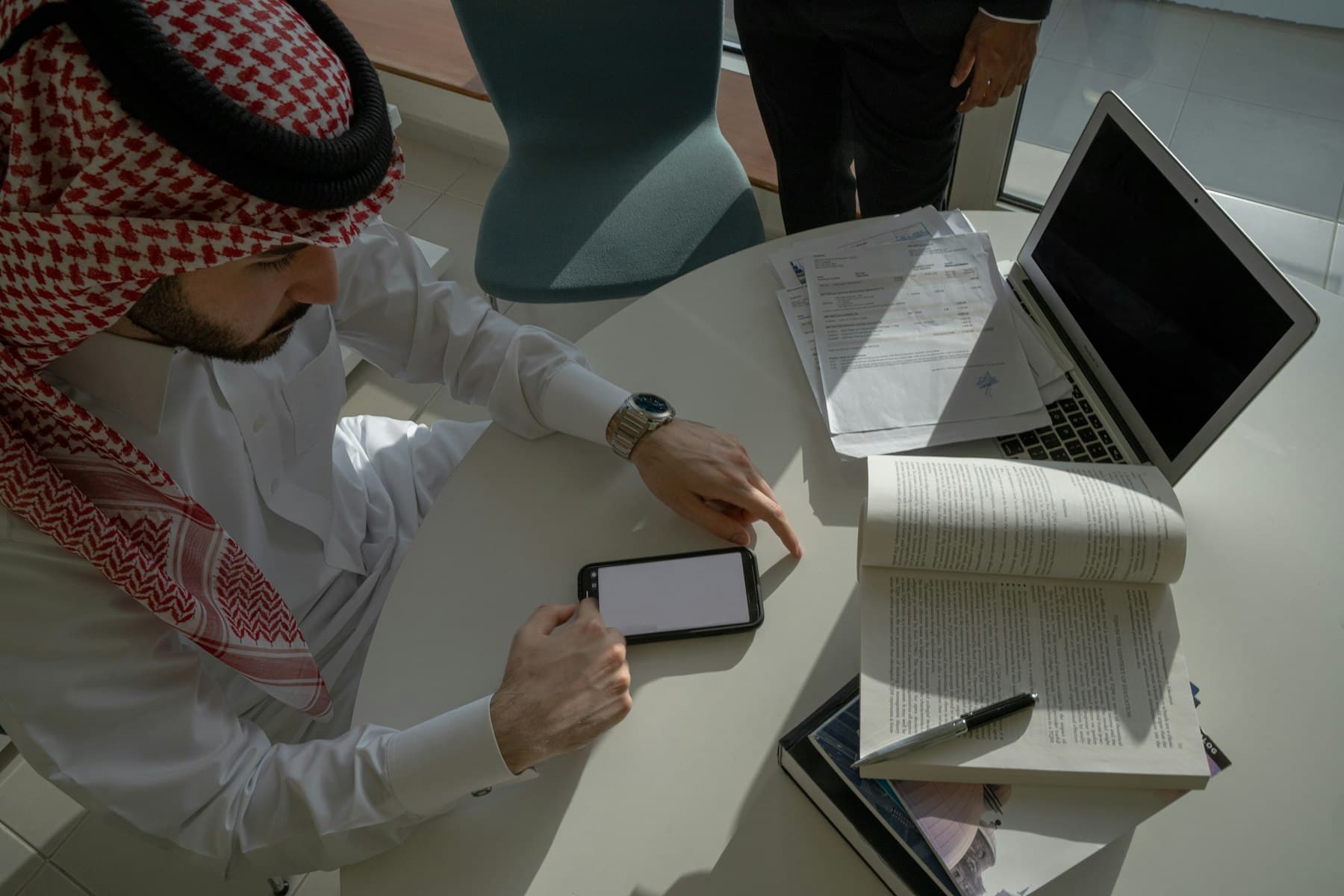 رجل أعمال بالزي القطري يخطّط على طاولة اجتماع وأمامه لوحٌ وأوراقٌ وحاسوب — لحظة تُجمع فيها الدراسة والتموضع والتقويم في خطةٍ واحدة